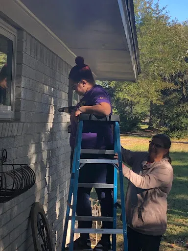 Two people steadying a ladder against a house on a bright afternoon.